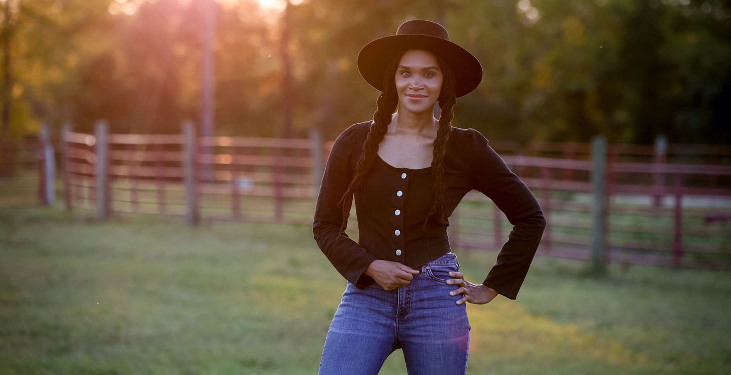 Una fotografía de una mujer vestida con una camisa negra, un sombrero de fieltro negro y unos vaqueros azules, posando en un prado verde.